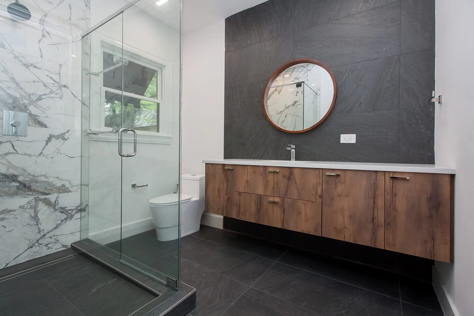 A bathroom with a marble shower, dark slate tile and a floating walnut vanity
