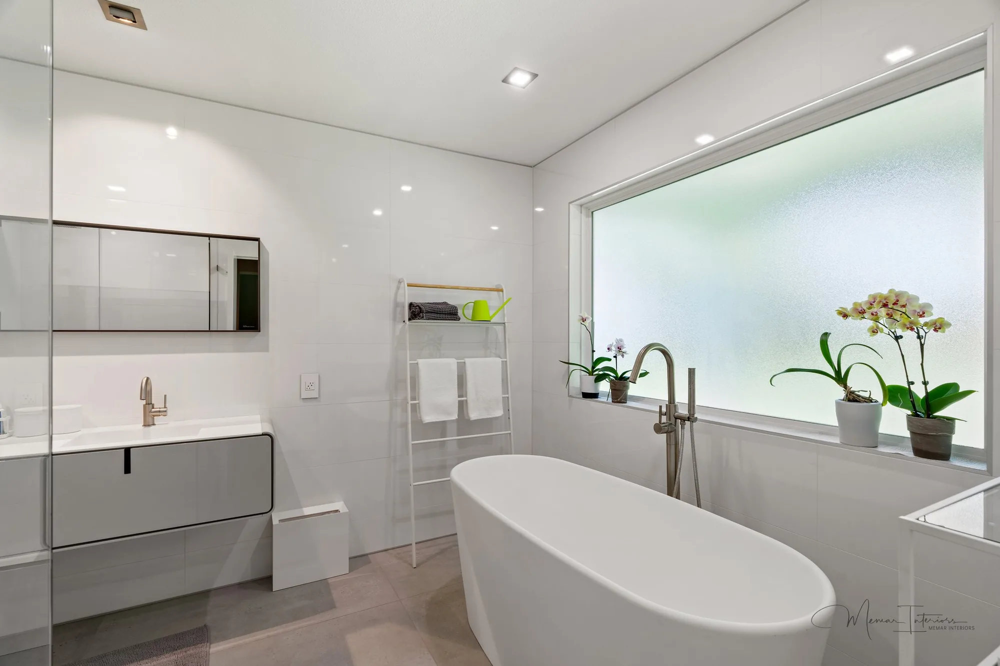 A white bathroom with a freestanding tub, a frosted glass window and orchids on the sill